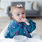A baby in a bow wears a Bumkins Sleeved Bib 2 Pack: Jungle and Animal Print, eating with a spoon from a gray bowl in a high chair.