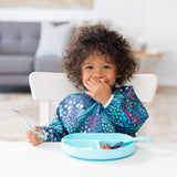 A toddler wearing a Bumkins Sleeved Bib 2 Pack: Jungle and Animal Print sits at a white table, covering their mouth with a blue plate nearby.