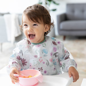 A girl wearing a floral bib in a high chair smiles as she self feeds from Bumkins Silicone First Feeding Set: Pink in a cozy living room.