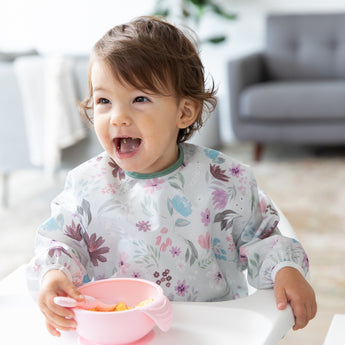 A girl wearing a floral bib in a high chair smiles as she self feeds from Bumkins Silicone First Feeding Set: Pink in a cozy living room.