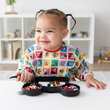 Smiling toddler with braided hair eats from a divided plate, wearing a colorful cartoon-themed bib at a white table.
