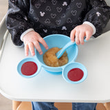 Close-up of a child in a Bumkins Sleeved Bib: Mickey Mouse Icon Black + White eating from a blue Mickey Mouse-shaped bowl.