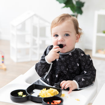 A boy in a high chair self feeds from a Mickey Mouse-shaped plate while wearing a Bumkins Sleeved Bib: Mickey Mouse Icon Black + White.