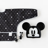 Bumkins Sleeved Bib: Mickey Mouse Icon Black + White lays flat next to a Mickey Mouse-shaped plate & matching utensils on a white background.