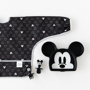 Bumkins Sleeved Bib: Mickey Mouse Icon Black + White lays flat next to a Mickey Mouse-shaped plate & matching utensils on a white background.