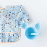 Bumkins Sleeved Bib: Mickey + Minnie Sunny Side Up, laid flat on a white background, is half visible next to a blue Mickey Mouse-shaped bowl and spoon set.
