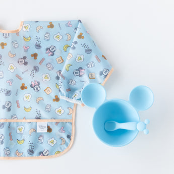Bumkins Sleeved Bib: Mickey + Minnie Sunny Side Up, laid flat on a white background, is half visible next to a blue Mickey Mouse-shaped bowl and spoon set.