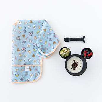 Bumkins Sleeved Bib: Mickey + Minnie Sunny Side Up is folded next to a Mickey-shaped bowl with yogurt and fruit and spoon on a white background.
