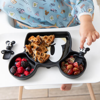 Close-up of a child's hand reaching for fruit & waffles from Bumkins Silicone Grip Dish: Mickey Mouse on a high chair tray.