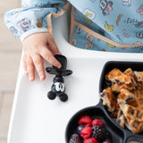 Close-up of a child's hand reaching for a Bumkins Silicone Chewtensils®: Mickey Mouse next to a plate of waffles & berries.