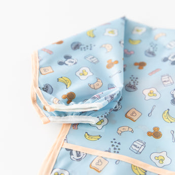 Close-up of Bumkins Sleeved Bib: Mickey + Minnie Sunny Side Up on a white background, showcasing the white elastic cuff on the sleeve.