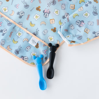 Two baby spoons, blue and black, sit beside a Bumkins Sleeved Bib: Mickey + Minnie Sunny Side Up on a white background.