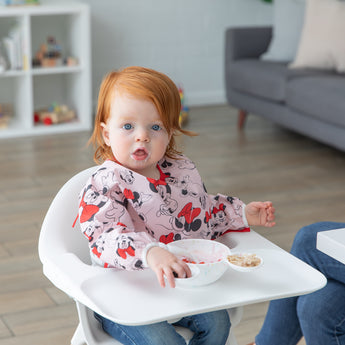 A young girl wearing a Bumkins Sleeved Bib: Minnie Mouse in a highchair eating from the Silicone First Feeding Set: Vanilla Sprinkle.