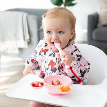 A young girl wearing a Bumkins Sleeved Bib: Minnie Mouse in a highchair eating from the Silicone First Feeding Set: Minnie Mouse Light Pink. 
