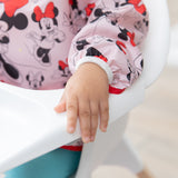 Close-up of a child with blue pants wearing a Bumkins Sleeved Bib: Minnie Mouse while sitting in a high chair.