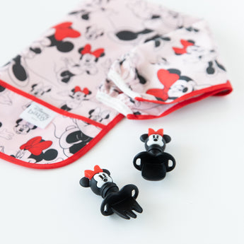 Bumkins Sleeved Bib: Minnie Mouse lays folded next to Minnie Mouse-shaped utensils on a white background.