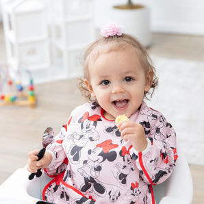 A young girl smiles, wearing a Bumkins Sleeved Bib: Minnie Mouse in a high chair while holding a spoon and a snack.