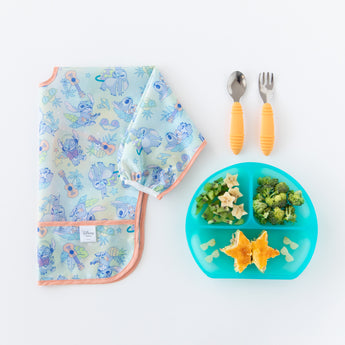 Bumkins Sleeved Bib: Aloha Stitch lays folded next to a blue divided plate with food and orange utensils on a white background.