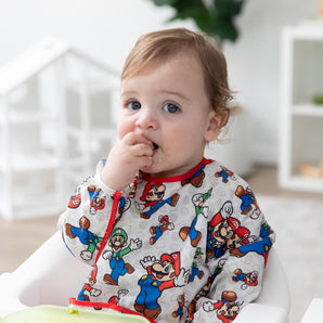 A light-haired boy wearing a Bumkins Sleeved Bib: Super Mario™ Classic in a high chair places food in their mouth.