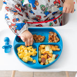 Close-up of a child in a colorful bib reaching for breakfast food from a blue plate next to a Bumkins Silicone Chewtensils®: Dark Blue.