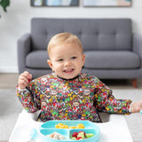 A boy wearing a Bumkins Sleeved Bib: Super Mario™ Mashup in a high chair smiles, excited to eat a meal from a blue plate on the tray.