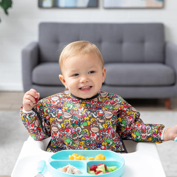 A boy wearing a Bumkins Sleeved Bib: Super Mario™ Mashup in a high chair smiles, excited to eat a meal from a blue plate on the tray.