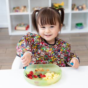 A smiling girl wearing a Bumkins Sleeved Bib: Super Mario™ Mashup eats breakfast from a green plate in front of toy-filled shelves.