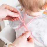 Adult hands tie the ribbon closure of a Bumkins Sleeved Bib 2 Pack: Disco Boots around a child's neck.