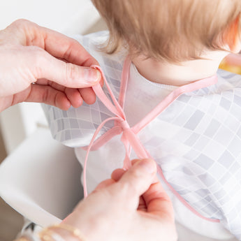 Adult hands tie the ribbon closure of a Bumkins Sleeved Bib 2 Pack: Disco Boots around a child's neck.
