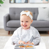 A smiling toddler in a Bumkins Sleeved Bib 2 Pack: Disco Boots sits in a high chair with food on a gray plate, wearing a white bow headband.