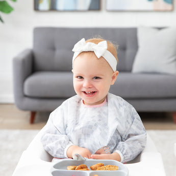 A smiling toddler in a Bumkins Sleeved Bib 2 Pack: Disco Boots sits in a high chair with food on a gray plate, wearing a white bow headband.