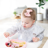 A toddler with a pink bow in a high chair wears a Bumkins Sleeved Bib 2 Pack: Disco Boots while eating from a pink plate with a fork.