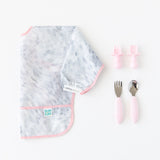 A folded Bumkins Sleeved Bib 2 Pack: Disco Boots rests next to two sets of pink toddler utensils on a white background.