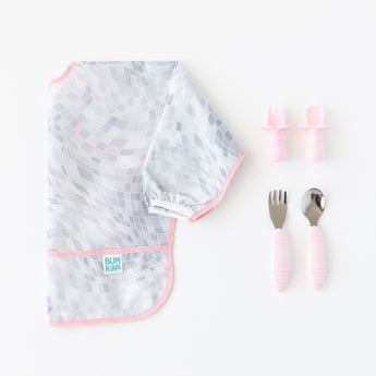 A folded Bumkins Sleeved Bib 2 Pack: Disco Boots rests next to two sets of pink toddler utensils on a white background.