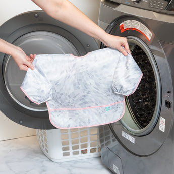 A person holds a Bumkins Sleeved Bib 2 Pack: Disco Boots in front of an open front-loading washing machine.