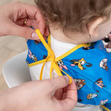 An adult ties the yellow ribbon closure of a Bumkins Sleeved Bib 2 Pack: Mickey & Friends Excellent Eight around a child's neck.