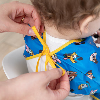 An adult ties the yellow ribbon closure of a Bumkins Sleeved Bib 2 Pack: Mickey & Friends Excellent Eight around a child's neck.