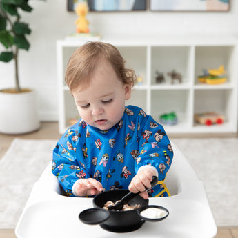 A toddler wears a Bumkins Sleeved Bib 2 Pack: Mickey & Friends Excellent Eight, sitting in a high chair eating from a black bowl with a spoon.
