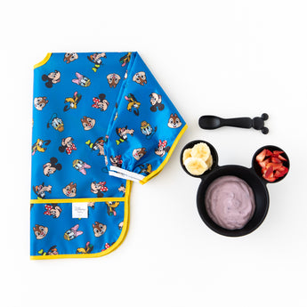 A Bumkins Sleeved Bib 2 Pack: Mickey & Friends Excellent Eight lays folded beside a bowl with yogurt & fruit and a spoon on a white background.
