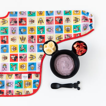 A bib from Bumkins Sleeved Bib 2 Pack: Mickey & Friends Excellent Eight lays flat next to a bowl & spoon set with yogurt & fruit on white.