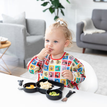 A toddler in a Bumkins Sleeved Bib 2 Pack: Mickey & Friends Excellent Eight eats from a Mickey Mouse-shaped plate at a white table.