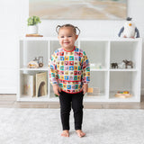 A smiling toddler wears a Bumkins Sleeved Bib 2 Pack: Mickey & Friends Excellent Eight, standing by a white shelf with toys.