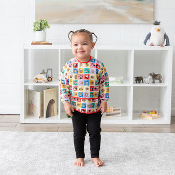 A smiling toddler wears a Bumkins Sleeved Bib 2 Pack: Mickey & Friends Excellent Eight, standing by a white shelf with toys.