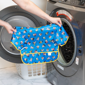 A person holds a Bumkins Sleeved Bib 2 Pack: Mickey & Friends Excellent Eight in front of an open front-loading washing machine.