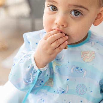 Close-up of a boy wearing a bib from Bumkins Sleeved Bib 2 Pack: Stitch Star-ring Up Trouble with his hand in his mouth.