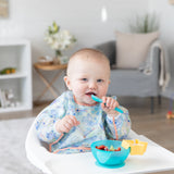 A smiling baby sits in a high chair, wearing a Bumkins Sleeved Bib 2 Pack: Stitch Star-ring Up Trouble while eating fruit from a blue bowl.