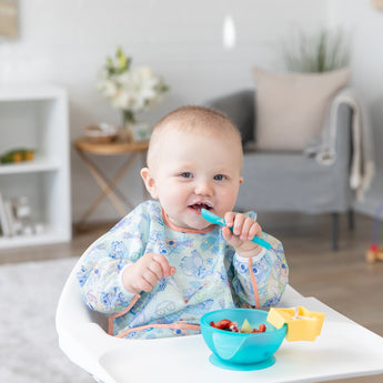 A smiling baby sits in a high chair, wearing a Bumkins Sleeved Bib 2 Pack: Stitch Star-ring Up Trouble while eating fruit from a blue bowl.