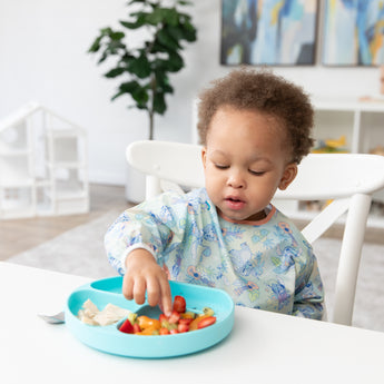 A toddler in a Bumkins Sleeved Bib 2 Pack: Stitch Star-ring Up Trouble picks up fruit from a blue plate at a white table in a bright room.