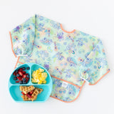 A bib from Bumkins Sleeved Bib 2 Pack: Stitch Star-ring Up Trouble lays flat next to a blue plate with breakfast food on a white background.
