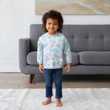A smiling child wears a Bumkins Sleeved Bib 2 Pack: Stitch Star-ring Up Trouble, standing barefoot on a rug in front of a gray sofa.